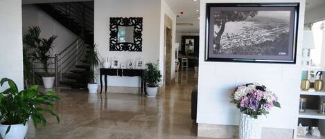 A modern lobby with a marble floor, a staircase, a reception desk, a large framed photograph, and potted plants.