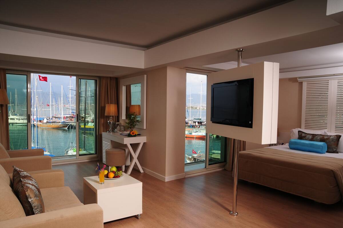 A hotel room with a bed, a TV, a desk, a chair, a coffee table with fruit, and large windows with a view of boats and a flag.