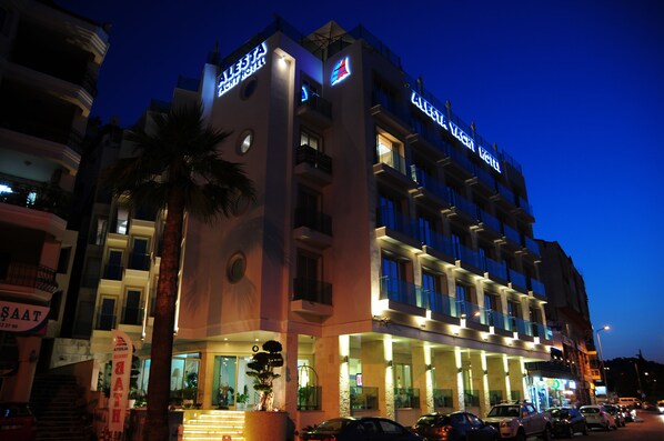 A multi-story hotel with illuminated signage, a palm tree in front, and a busy street with parked cars. - Alesta Yacht Hotel (Fethiye)