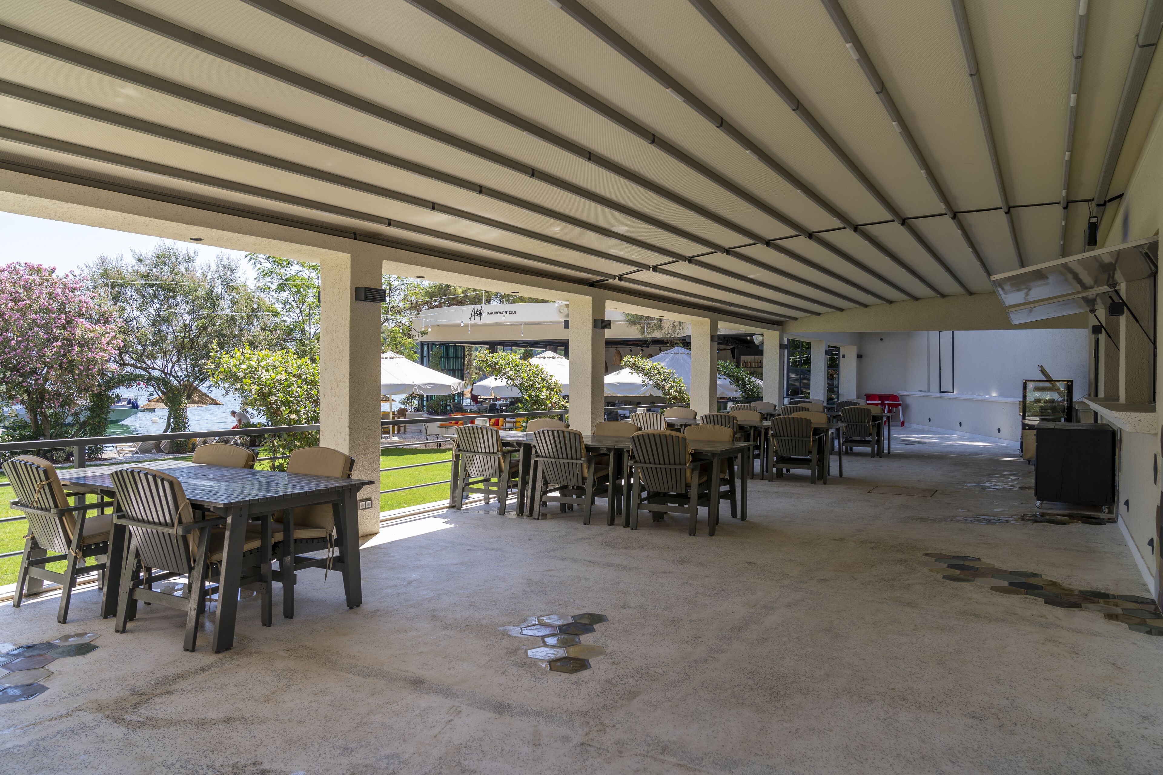 An outdoor patio area with tables and chairs, a covered walkway, and a view of the surrounding area including trees and a building.