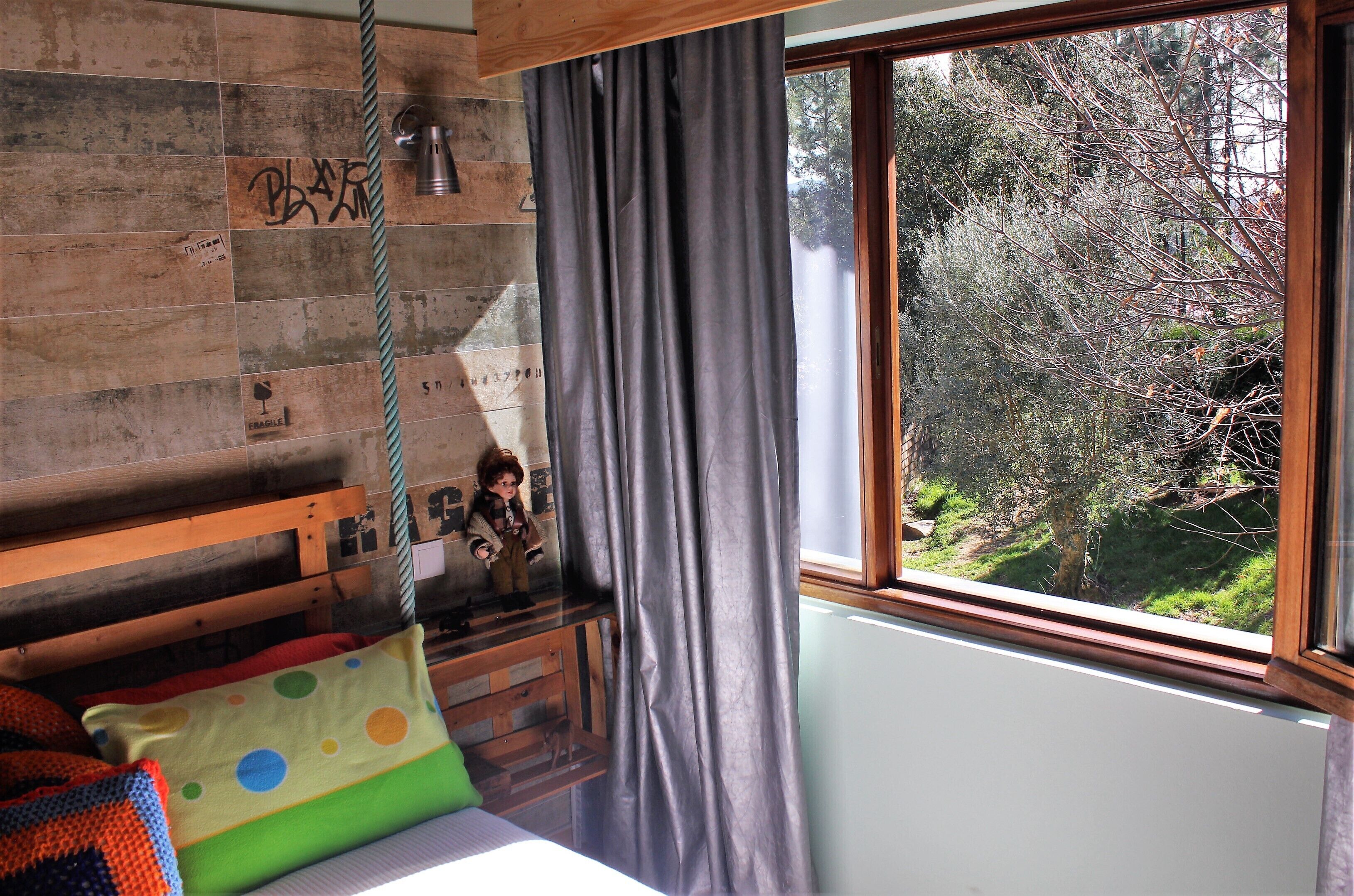 Quarto de criança com cabeceira de madeira, janela com vista para árvores e um balanço pendurado.