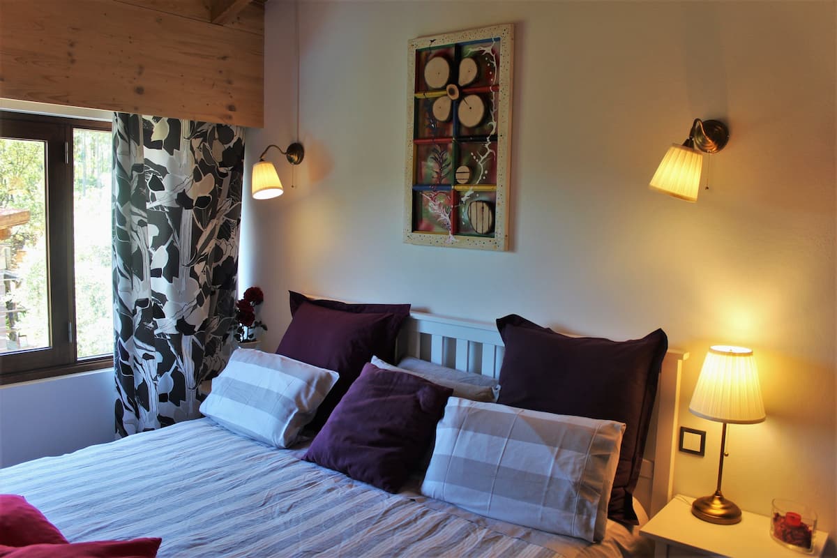 Ein Schlafzimmer mit einem Bett, Kissen, einem Nachttisch, einer Lampe, einem Wandbild und einem Fenster mit Vorhängen.
