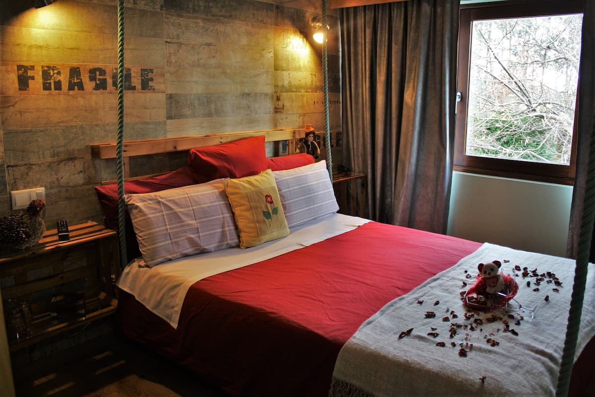 Ein Bett mit roter Bettwäsche, ein Holz-Kopfteil und ein Fenster mit Blick auf Bäume.