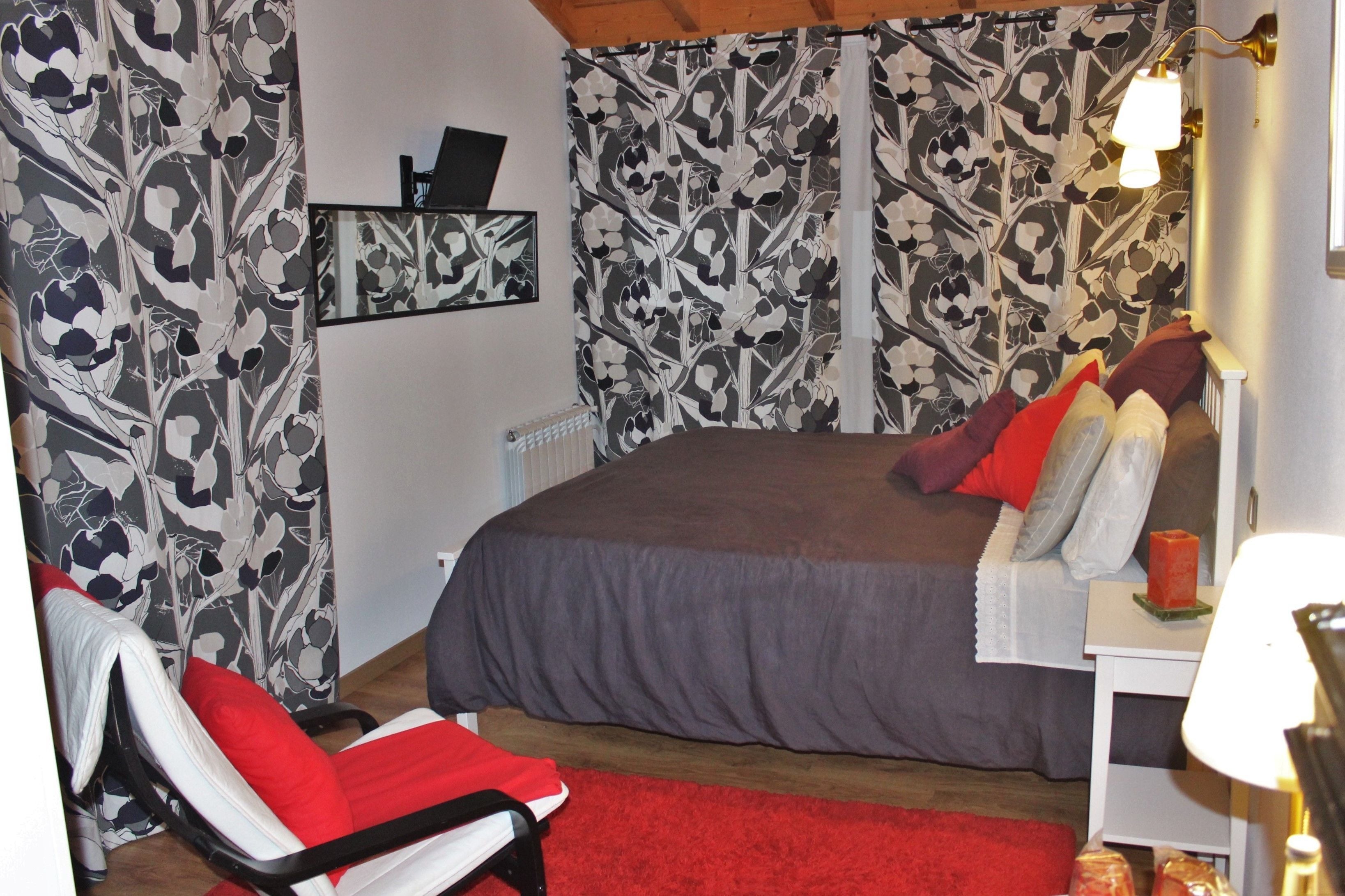Un dormitorio con una cama grande, una silla roja, una mesita con una lámpara y un papel tapiz estampado.