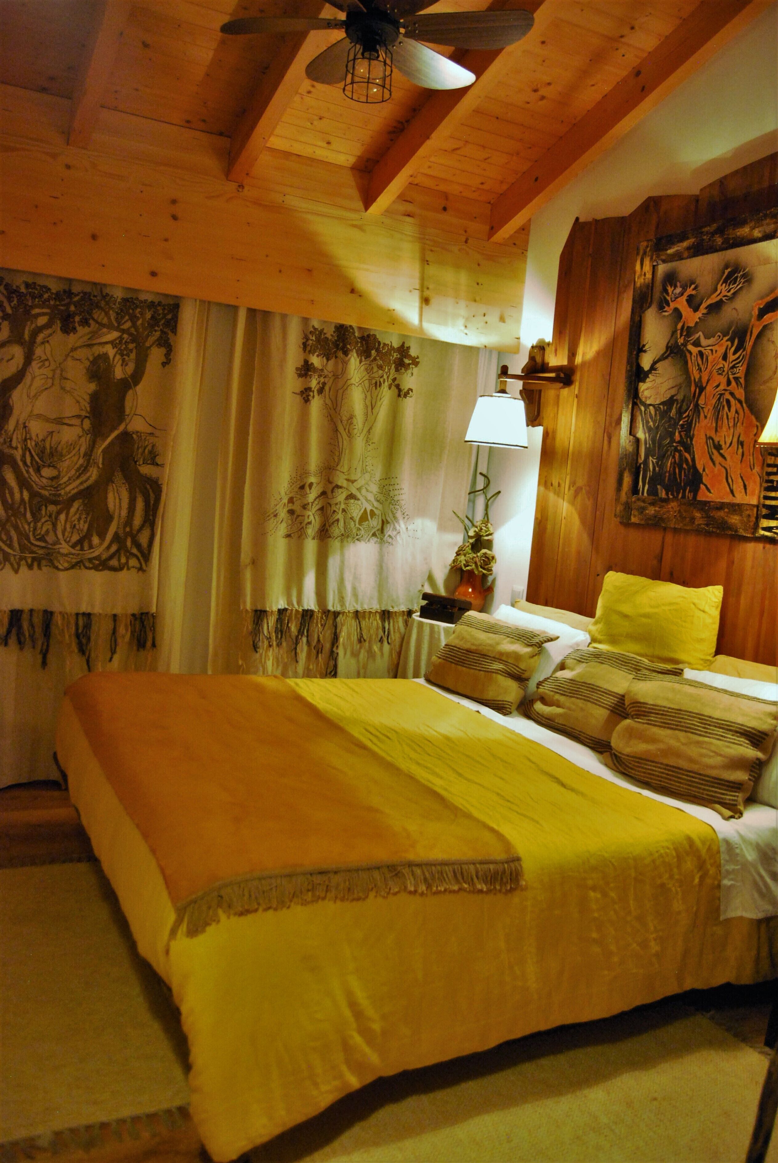 A room with a bed, wooden ceiling, a painting, curtains, and a lamp.