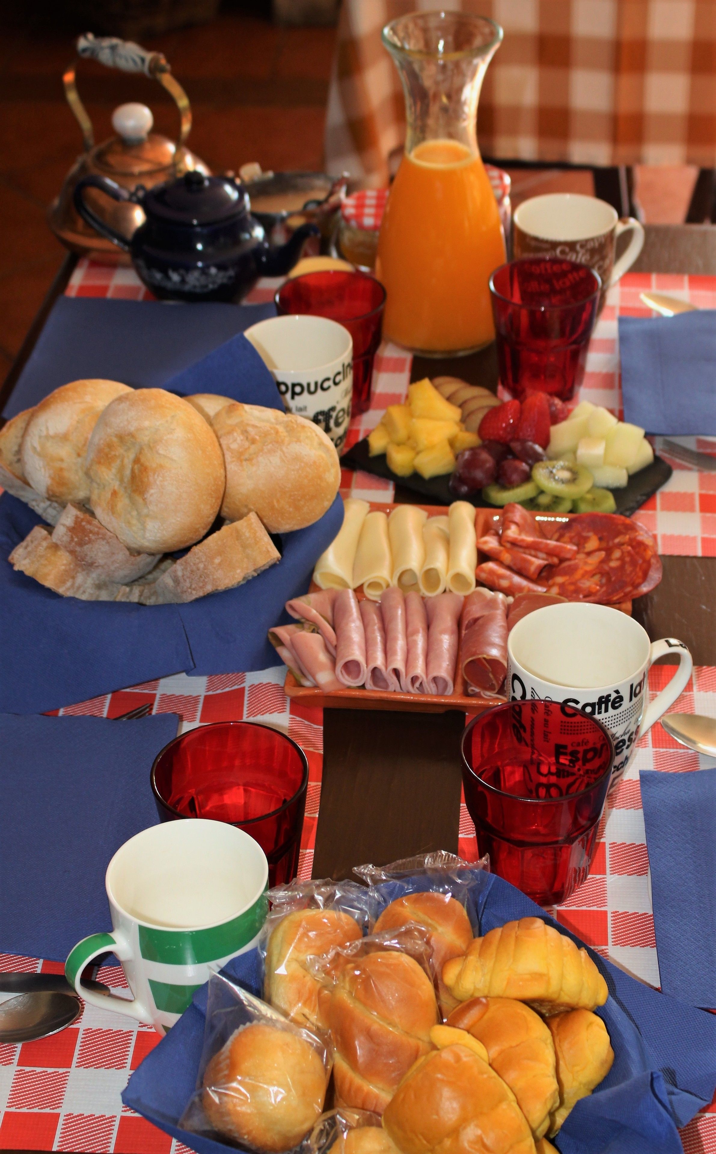 Una mesa de desayuno con pan, bollería, queso, embutidos y una jarra de zumo de naranja.