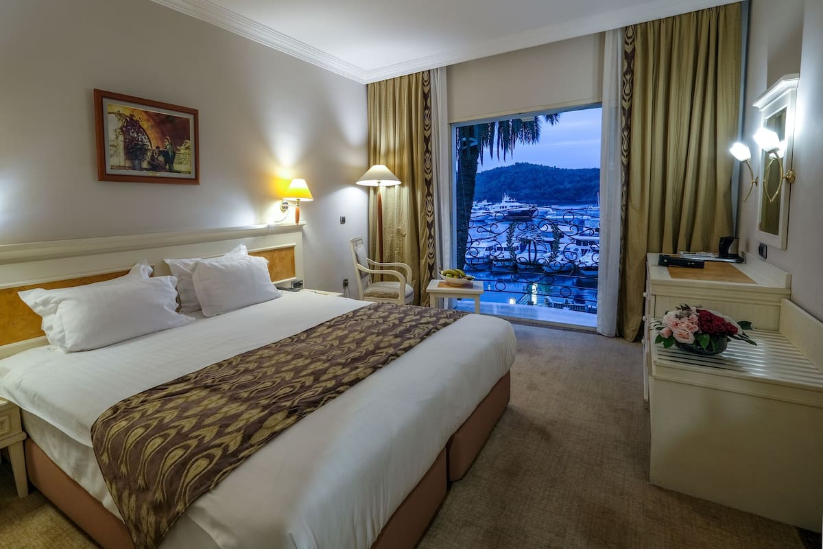 Quarto de hotel com uma cama grande, duas luminárias de cabeceira, uma cadeira, uma escrivaninha com luminária, um espelho e vista para a paisagem urbana pela janela.
