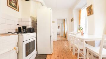 A kitchen with white appliances, a stove, and a refrigerator. There is a dining table with chairs and a window with curtains.