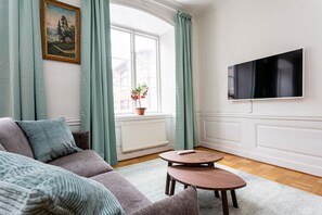 A living room with a sofa, two round tables, a TV mounted on the wall, a window with curtains, and a potted plant on the windowsill. - ApartDirect Gamla Stan II (Stockholm)