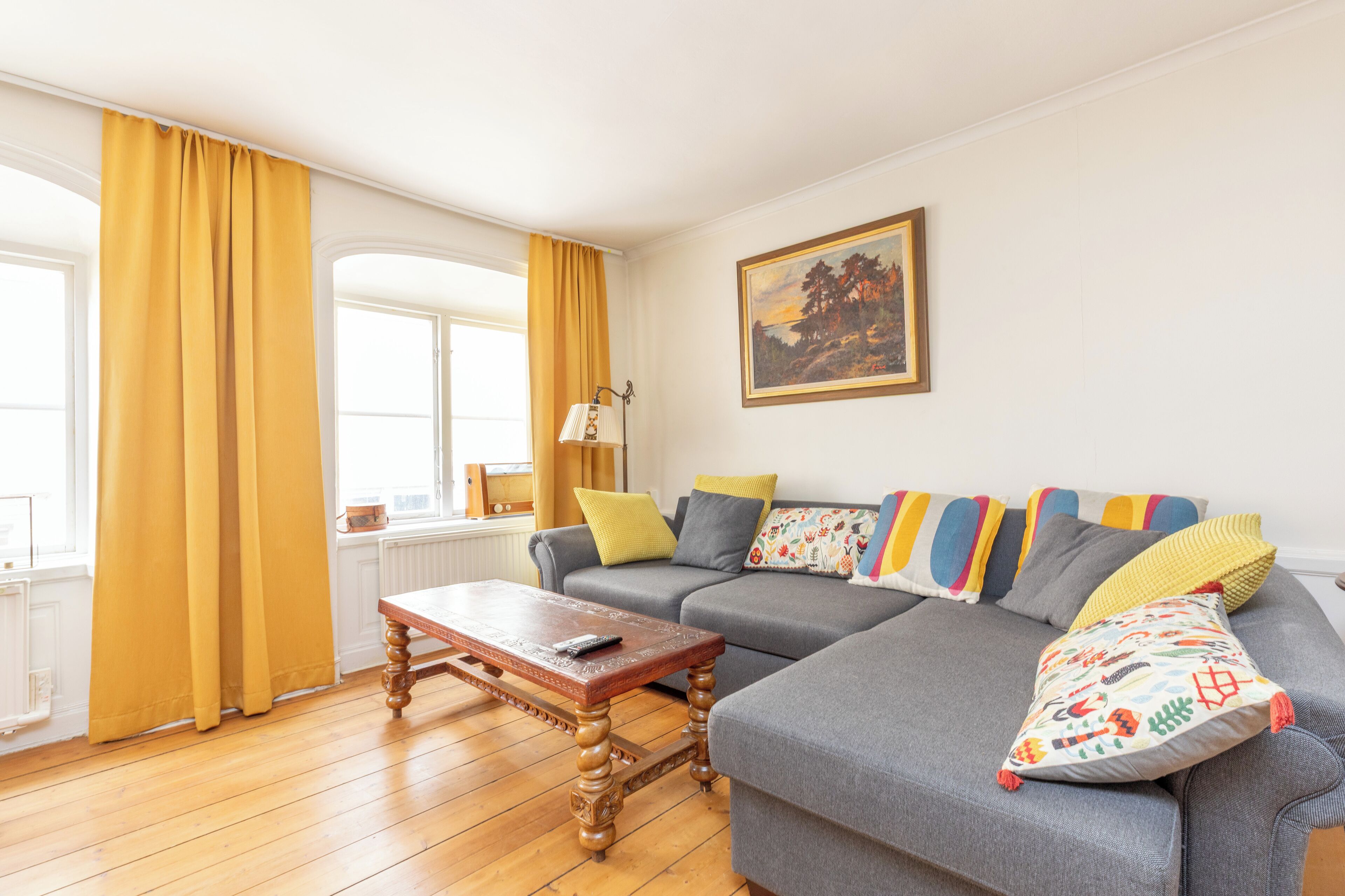 Una sala de estar con un sofá, una mesa de centro de madera, cortinas amarillas y un cuadro en la pared.