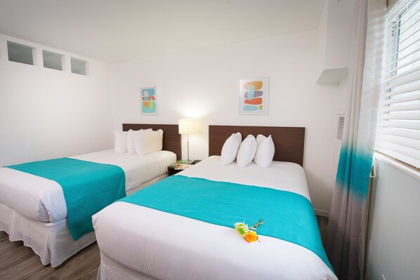 Habitación de hotel con dos camas, cada una con ropa de cama blanca y detalles en color aguamarina. Hay apliques de luz y obras de arte sobre las camas.