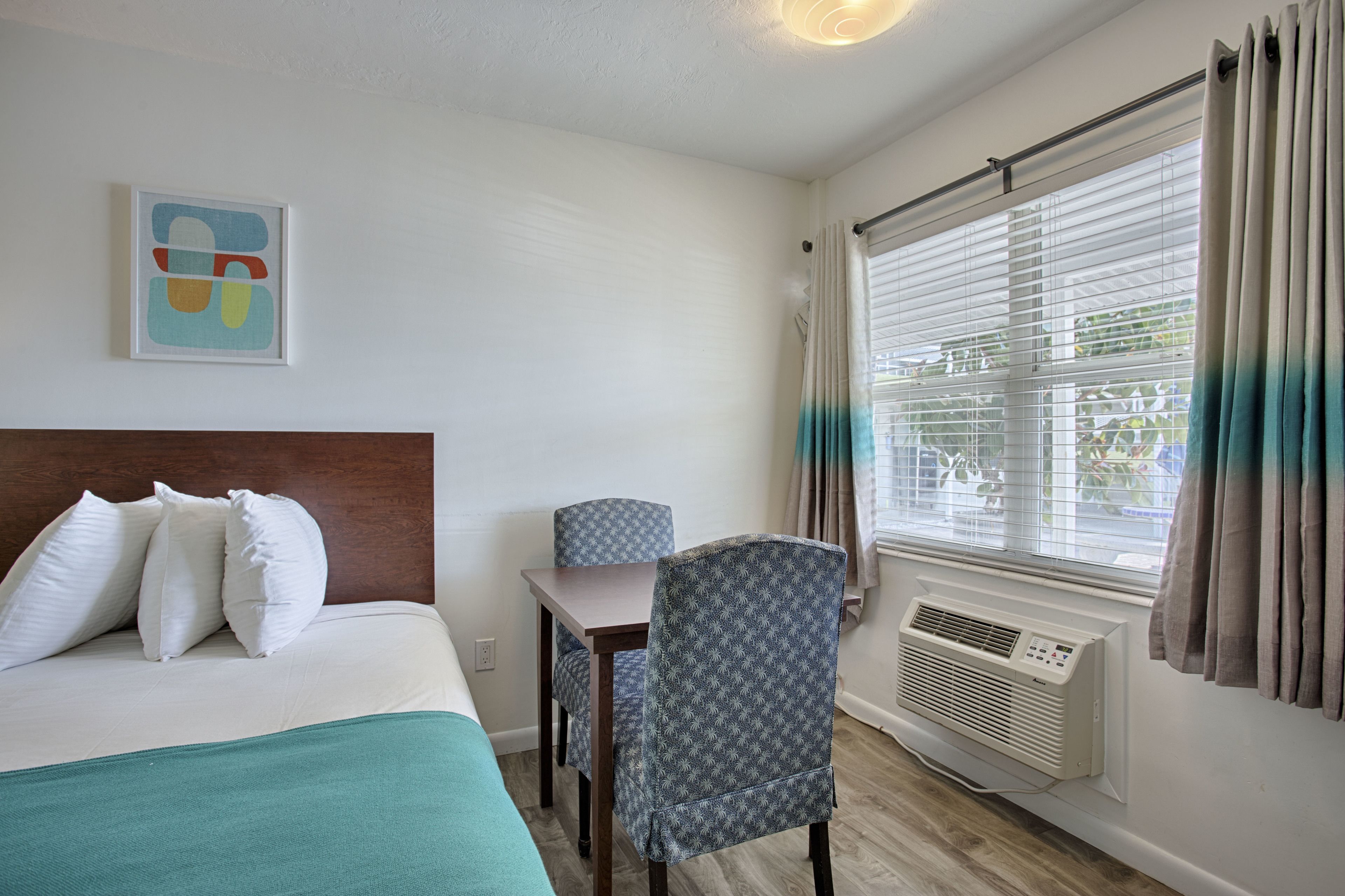 Una habitación de hotel con cama, escritorio, silla, ventana con cortinas y aire acondicionado.