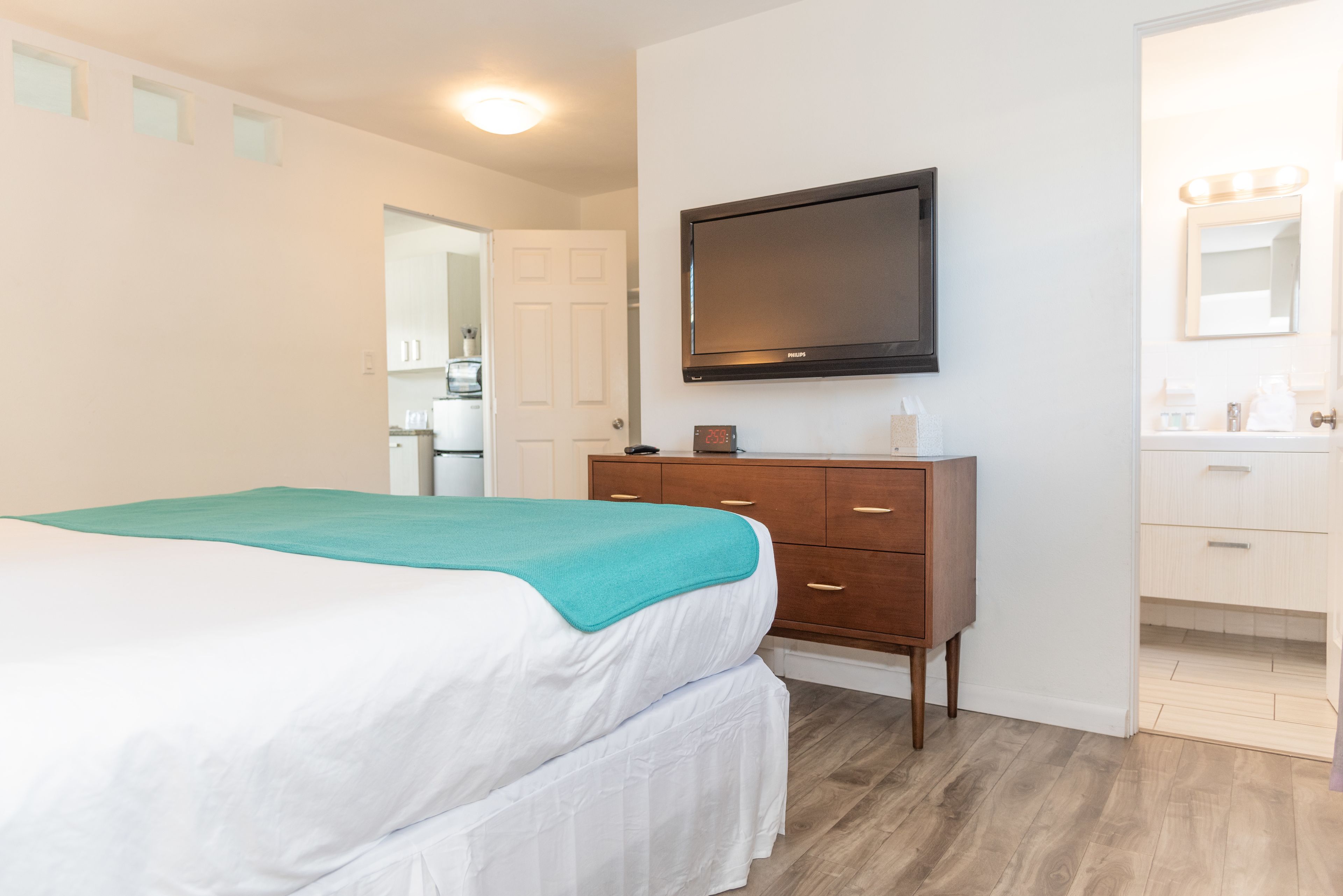 Une chambre d’hôtel avec un lit, une commode en bois, un téléviseur fixé au mur et une salle de bain visible à travers une porte ouverte.