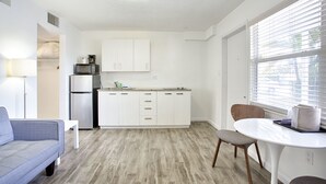Una cocina moderna con gabinetes blancos, microondas integrado y refrigerador. Hay una mesa de comedor con sillas y una ventana con persianas.