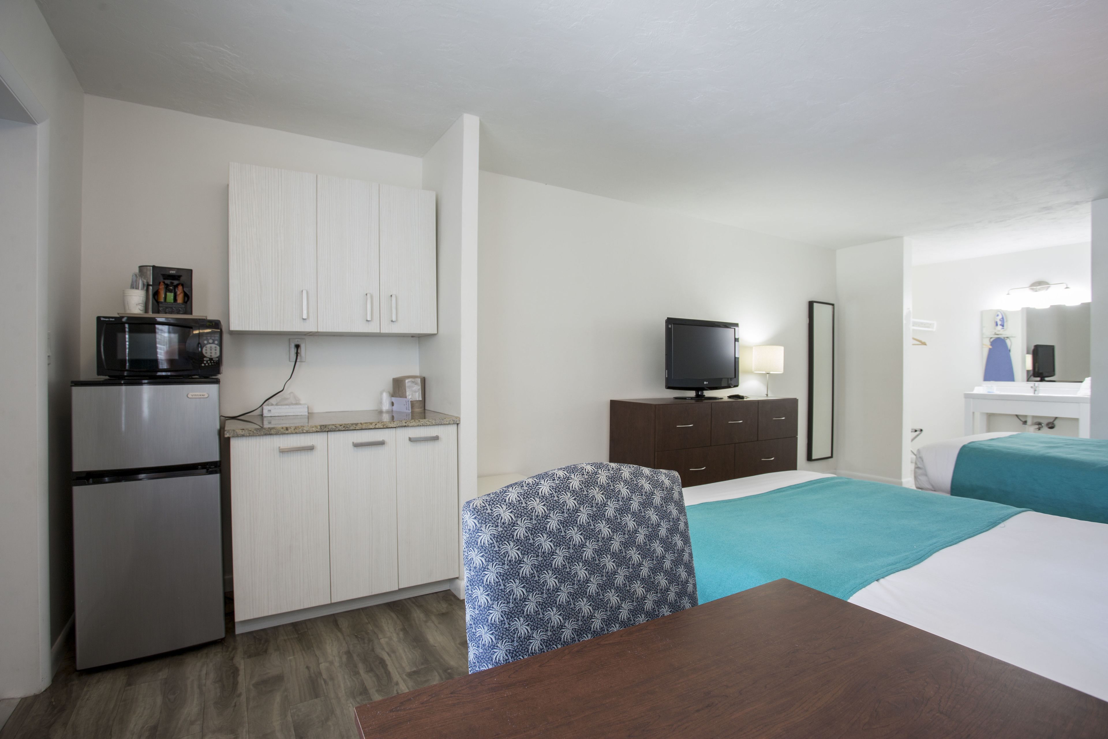 A compact hotel room with a kitchenette, a bed with teal bedding, a wooden table, and a patterned chair.