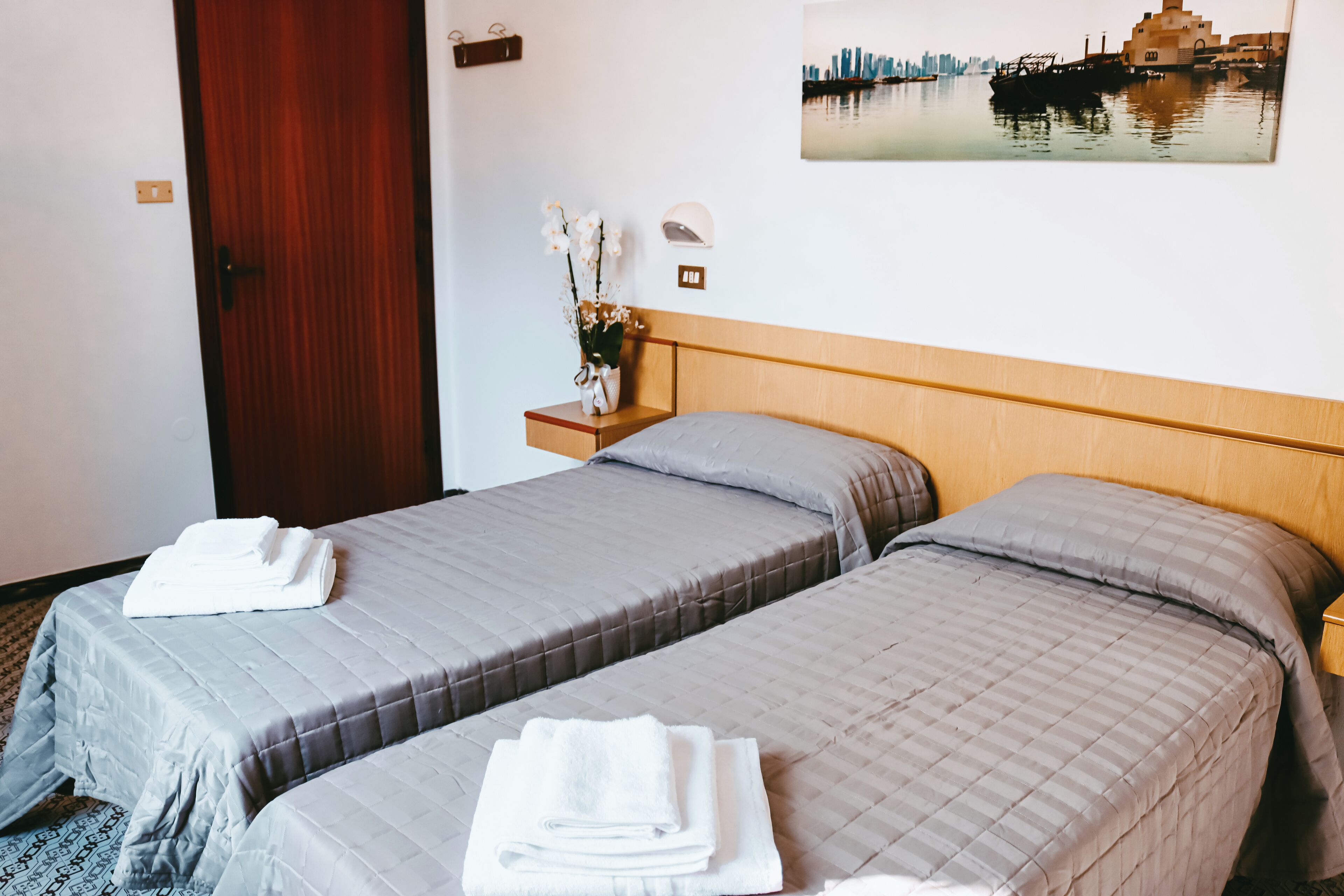 A hotel room with two beds, a wooden headboard, a nightstand with a vase of flowers, and a cityscape painting on the wall.