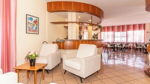 Un hall d’hôtel avec deux fauteuils blancs, une table en bois ornée d’un vase de fleurs, un comptoir d’accueil et un espace repas avec des chaises et des tables.