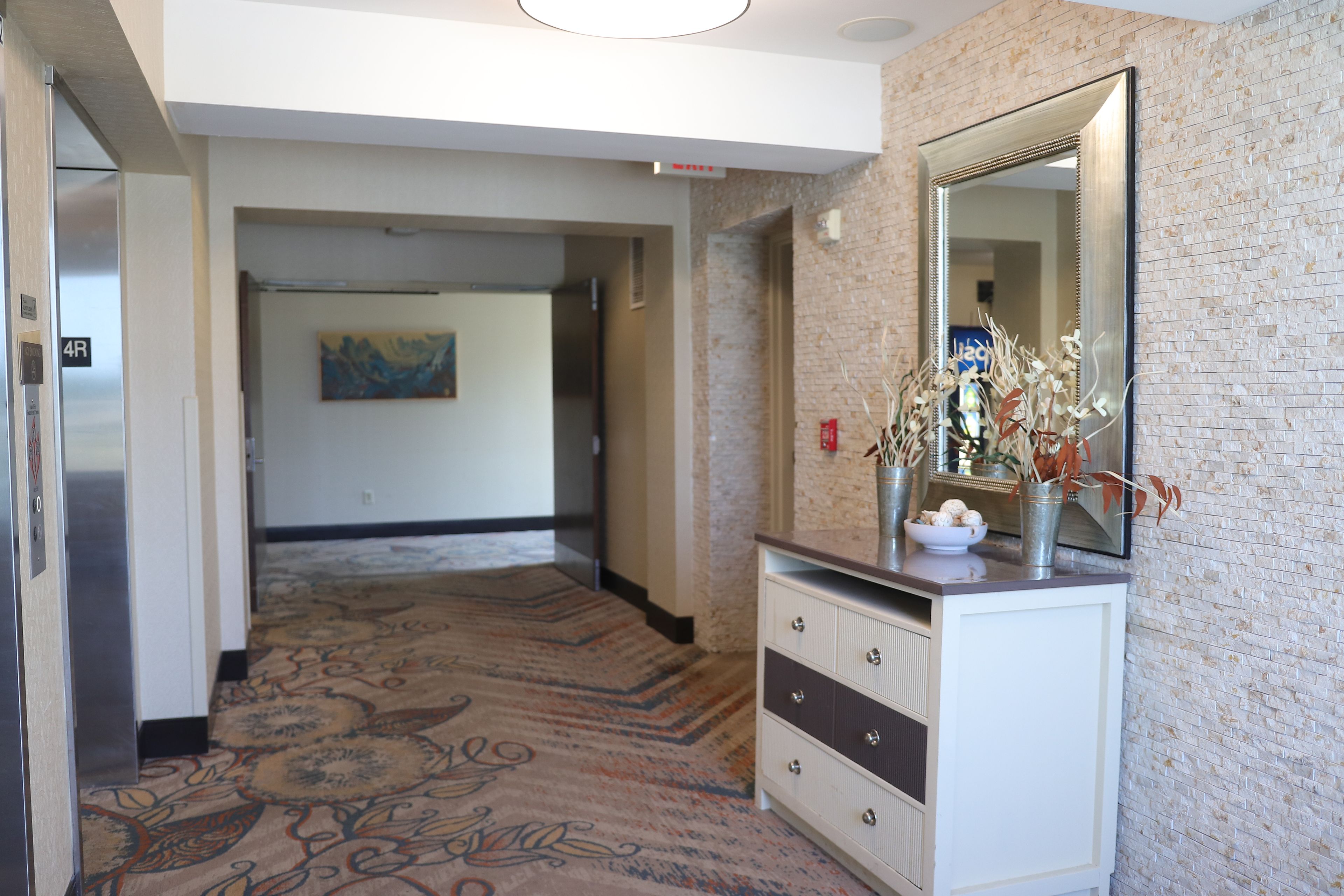 A hotel hallway with a carpeted floor, a dresser with a mirror, and a painting on the wall.