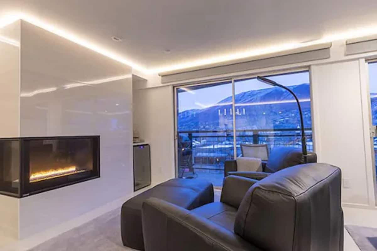 Un salon moderne doté d’une cheminée, d’un canapé et d’une grande fenêtre offrant une vue sur le paysage urbain et les montagnes.