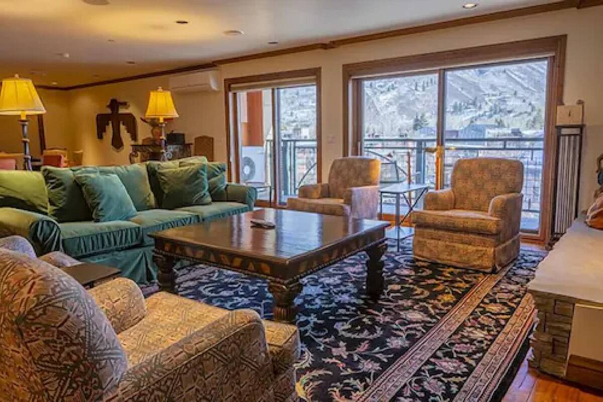 Un salon chaleureux avec un canapé vert, des fauteuils à motifs, une table basse et une grande fenêtre donnant sur une vue imprenable sur les montagnes.