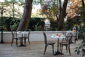 An outdoor dining area with wooden tables and chairs, surrounded by trees and greenery.