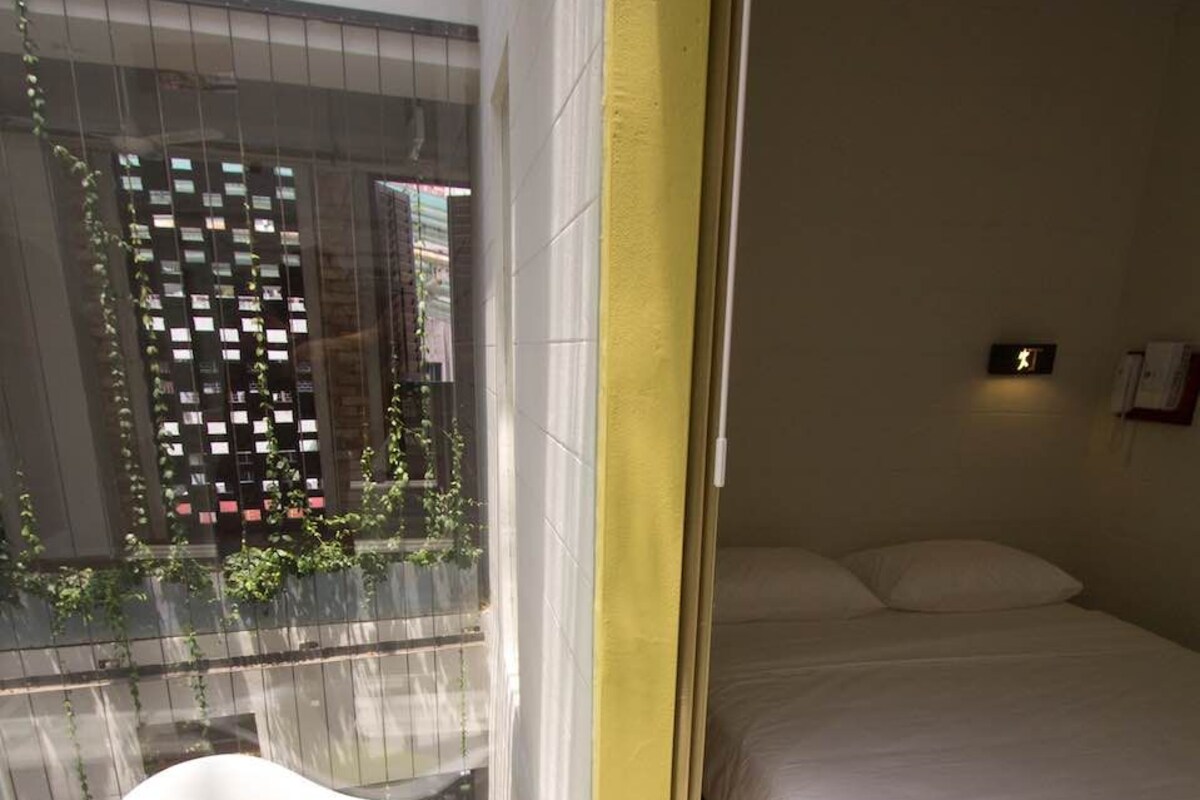 Quarto moderno com uma cama, uma cadeira branca com assento verde e vista para edifícios pela janela.