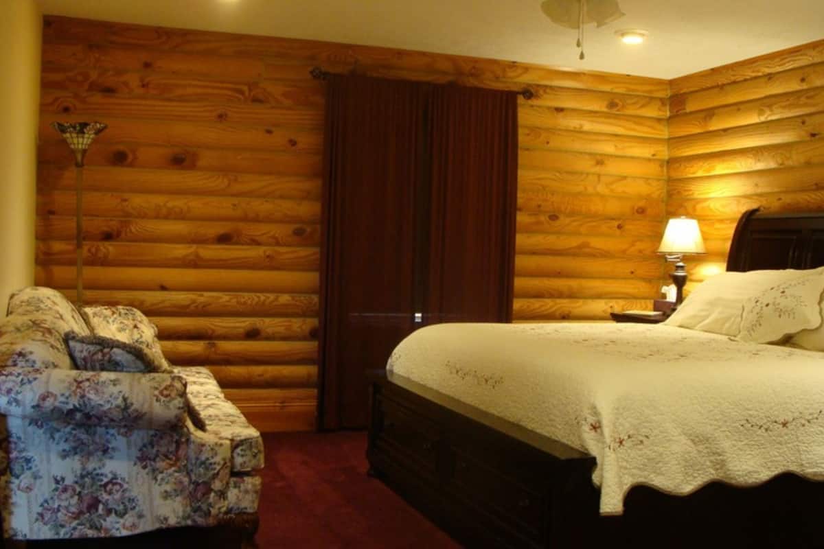 A log cabin-style bedroom with a wooden wall, a bed with a white bedspread, a nightstand with a lamp, and a floral-patterned armchair.