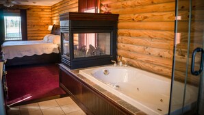 A log cabin-style bedroom with a large bed, a glass-enclosed bathtub, and a fireplace. - A Breath of Heaven B&B (Traverse City)
