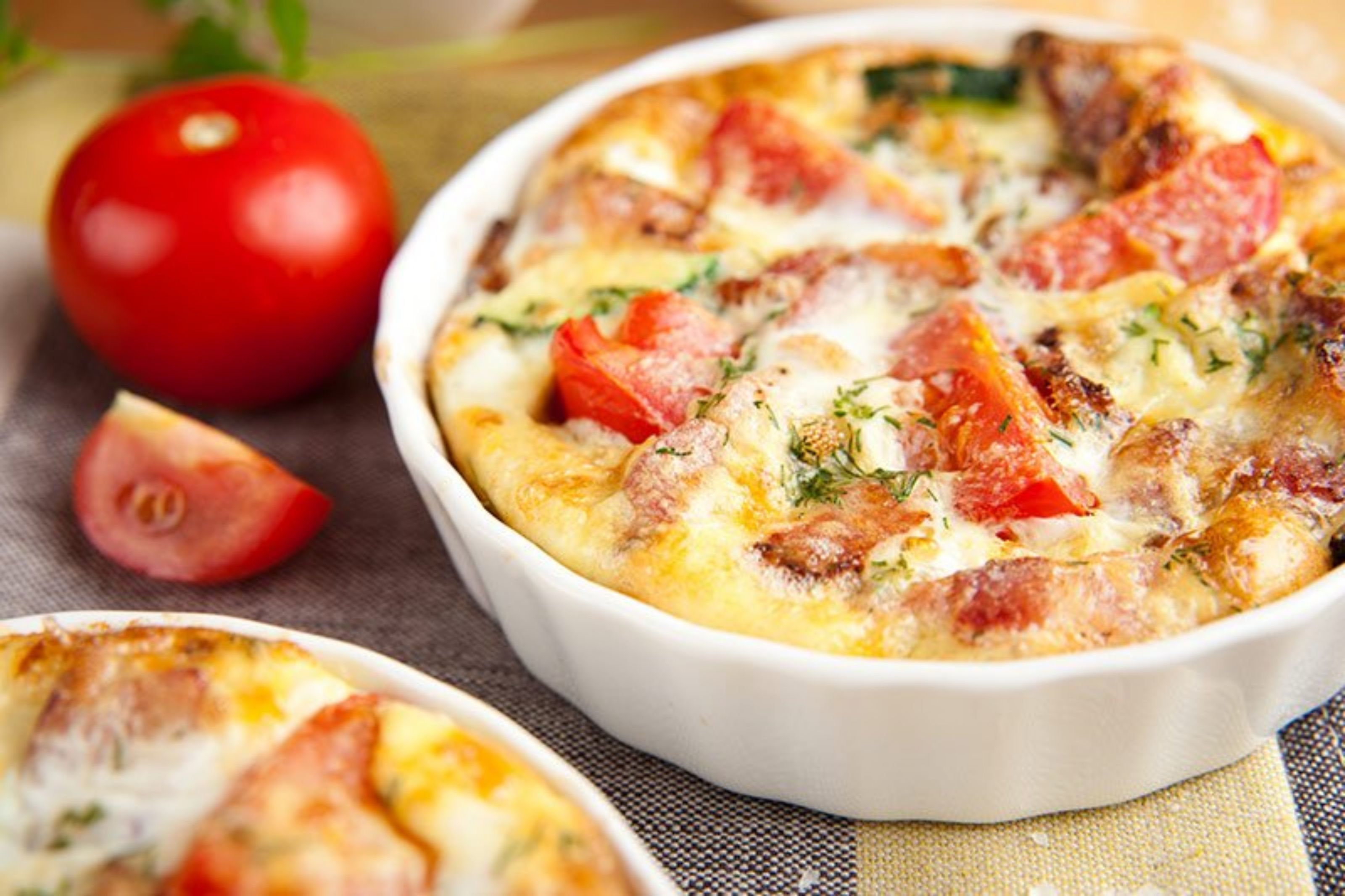 Un plato blanco de cerámica con un guiso al horno que lleva queso, tomates y hierbas.