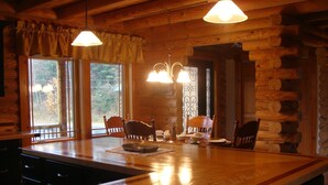A rustic wooden interior with a large island table, two hanging pendant lights, and a view through the windows. - A Breath of Heaven B&B (Traverse City)