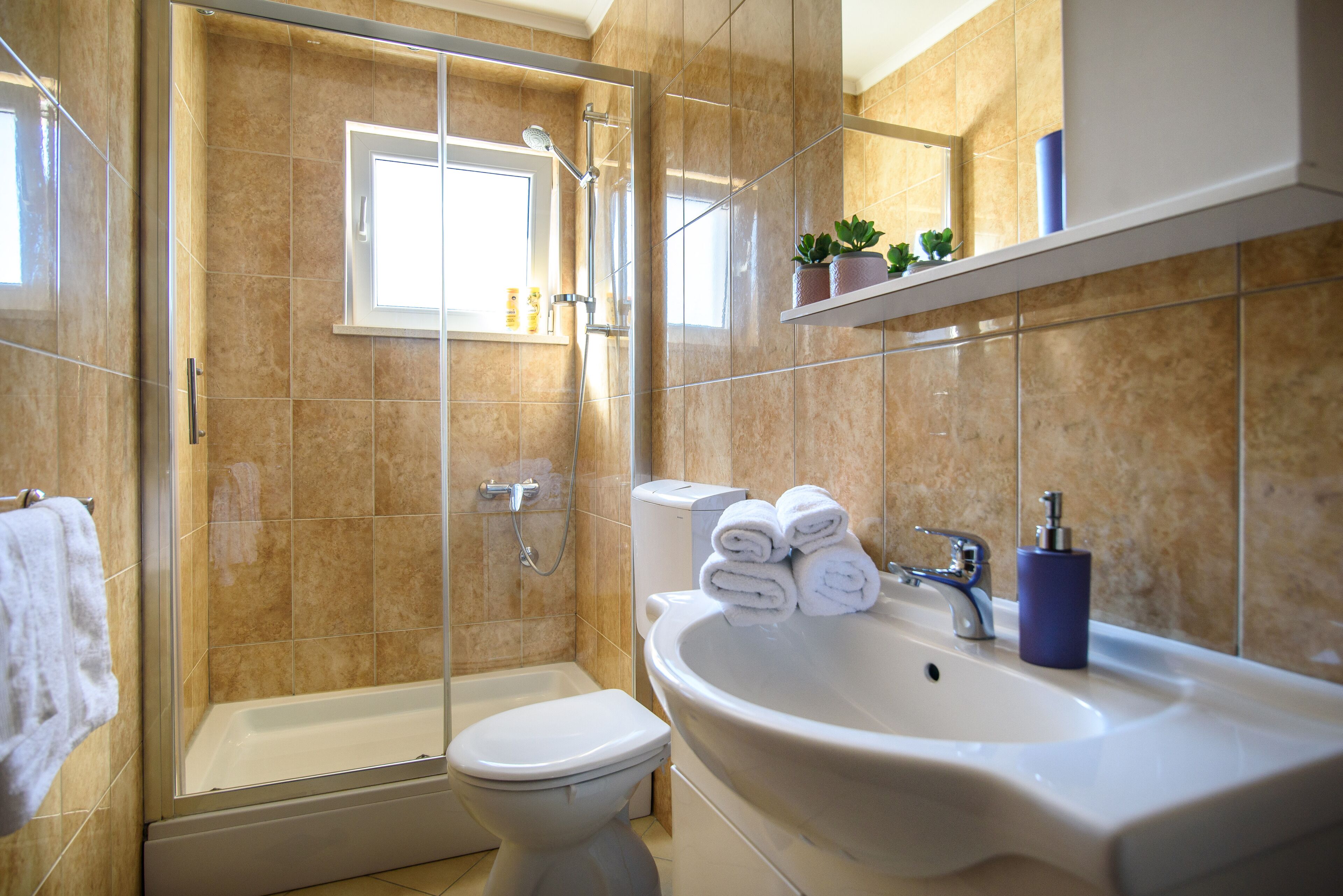 A bathroom with a glass-enclosed shower, a white toilet, a sink with a blue soap dispenser, and a window above the toilet.