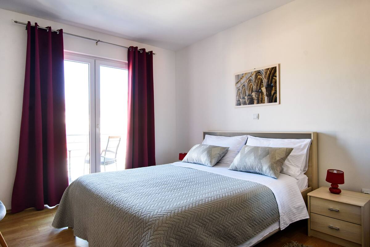 Quarto com cama, mesa de cabeceira, abajur vermelho, pintura na parede e vista pela janela.