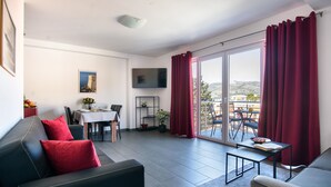 A modern living room with a black leather sofa, red cushions, a dining table with chairs, a flat-screen TV, and a balcony with red curtains.