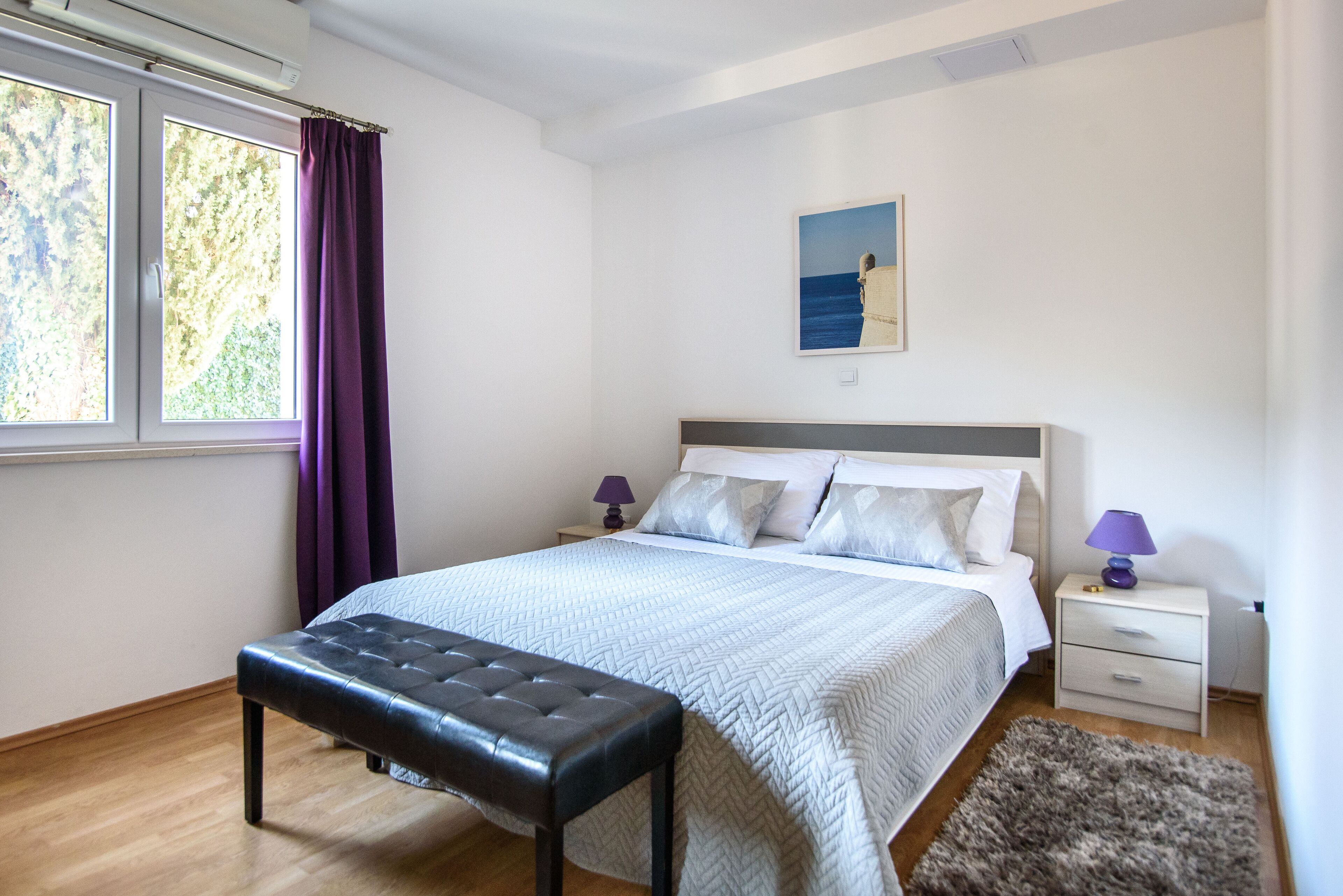 Een slaapkamer met een bed, twee nachtkastjes, een bankje, een raam met gordijnen en een schilderij aan de muur.