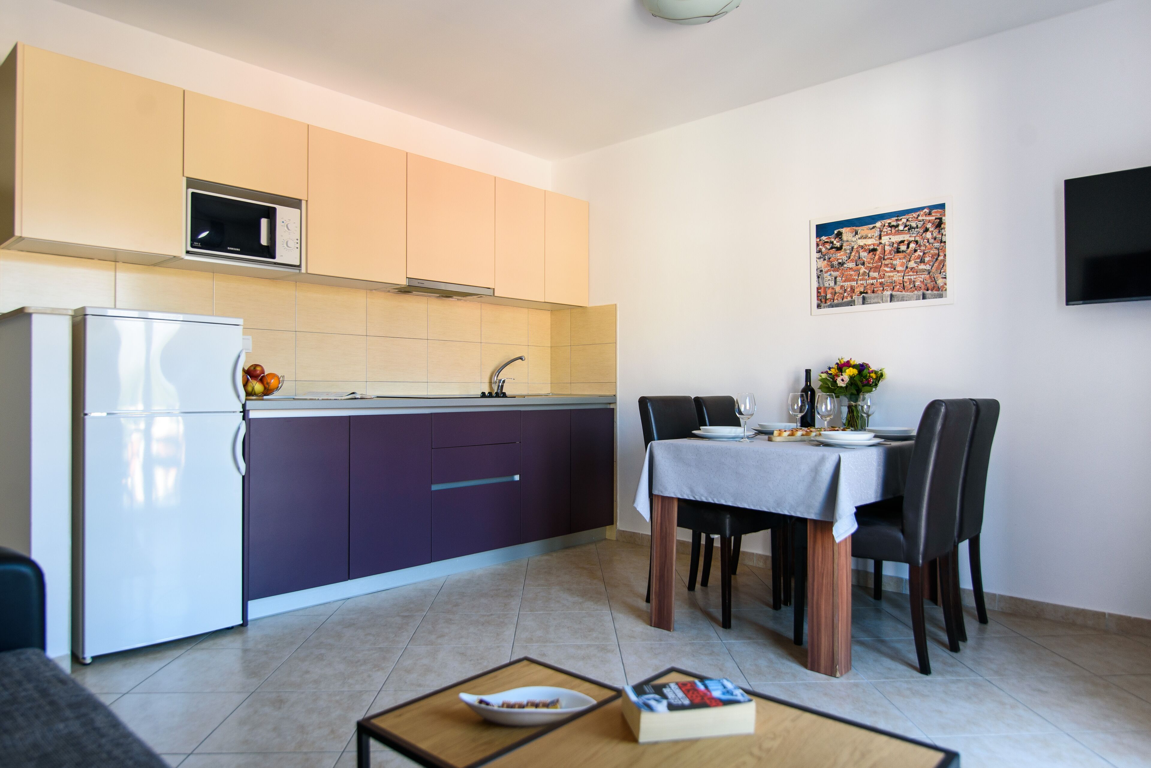A modern kitchen with a refrigerator, microwave, and purple cabinets. There is a dining table with chairs and a vase of flowers.