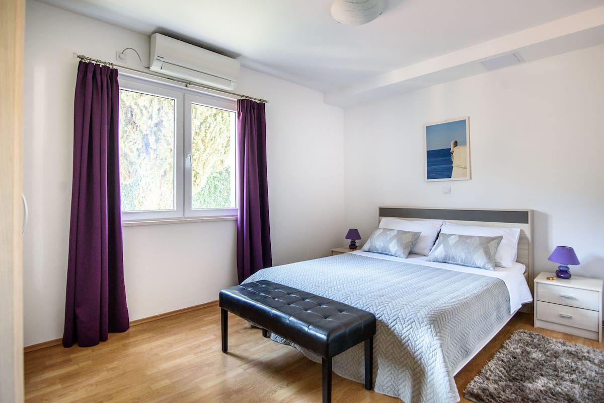 Quarto com cama, um banquinho, mesa de cabeceira e janela com cortinas.