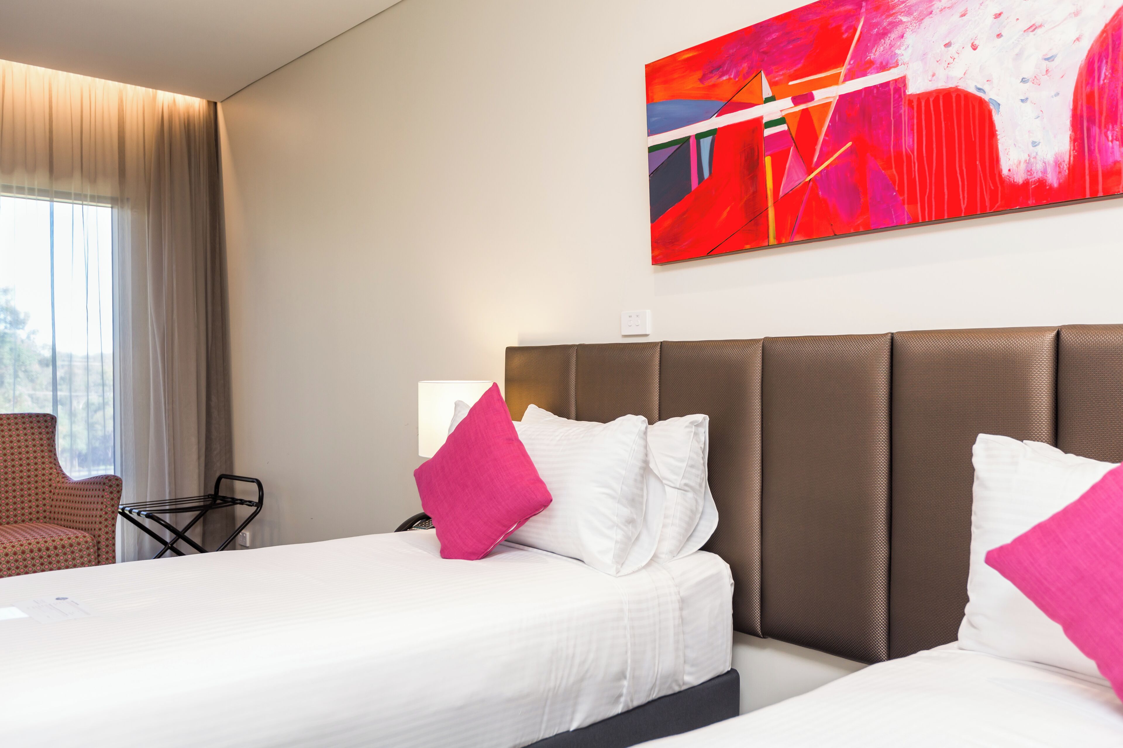 Une chambre d’hôtel avec deux lits, une grande peinture abstraite, une fenêtre avec des rideaux et une chaise.