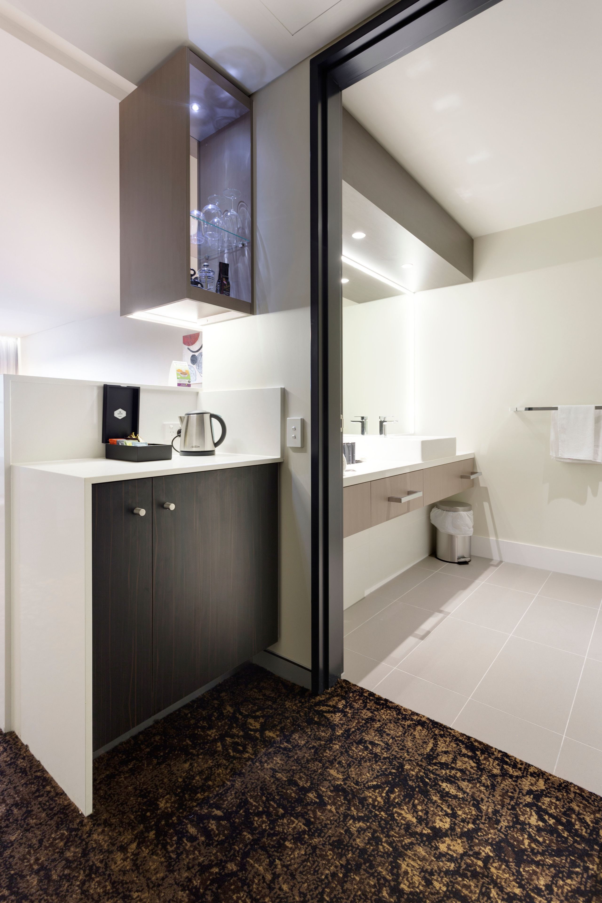 A modern bathroom with a dark wood cabinet, a coffee maker, and a glass shelf with bottles.