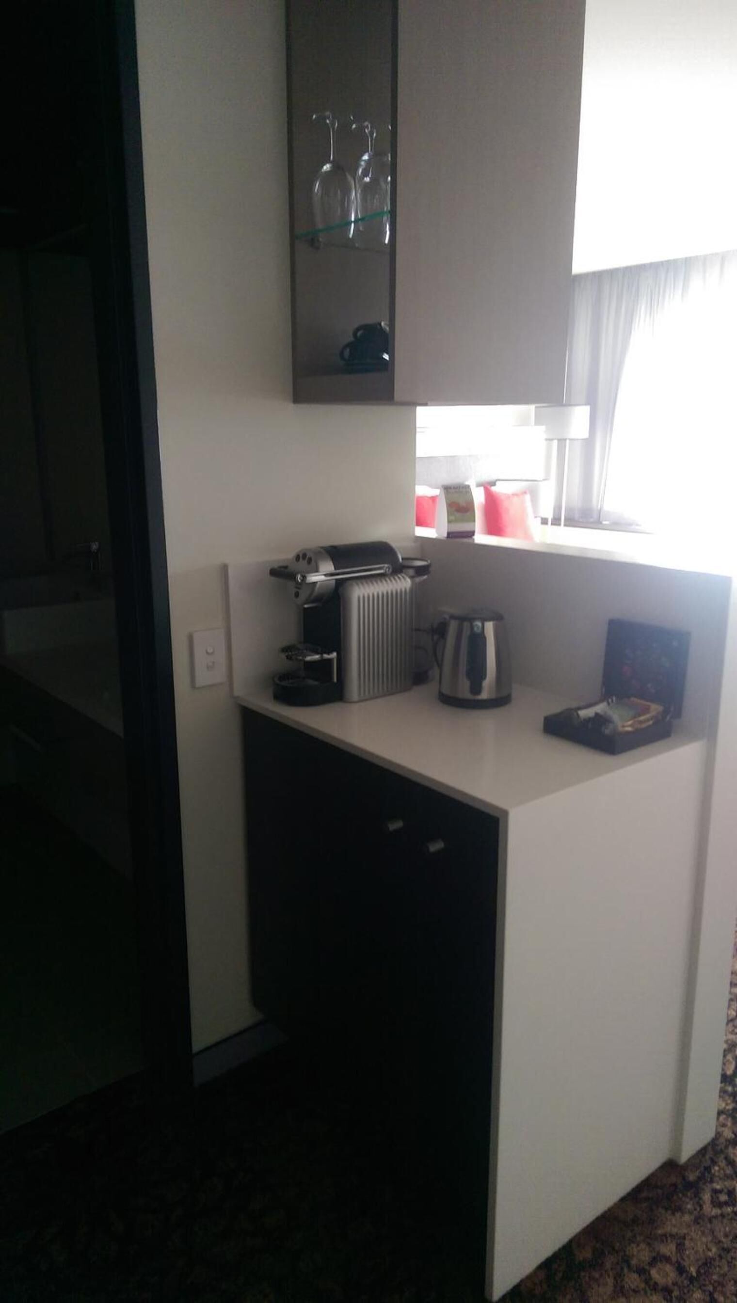 Kompakti hotellihuone, jossa on pieni keittiönurkkaus, pieni jääkaappi, kahvinkeitin ja ikkuna, jossa on verhot.