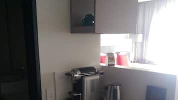 Kompakti hotellihuone, jossa on pieni keittiönurkkaus, pieni jääkaappi, kahvinkeitin ja ikkuna, jossa on verhot.