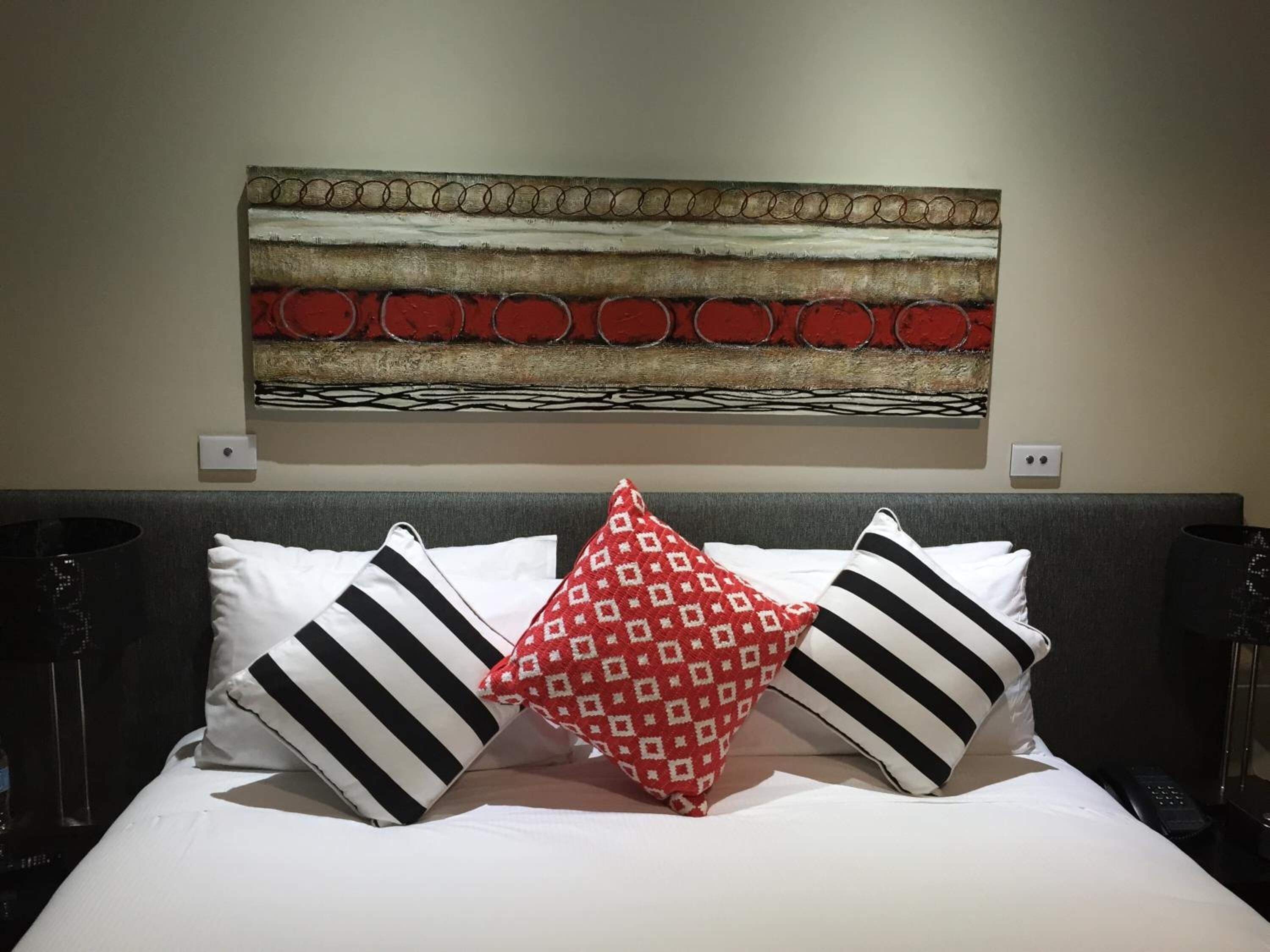 Una cama bien tendida con almohadas a rayas y estampadas, un adorno en la pared superior y una pared lisa con dos interruptores.