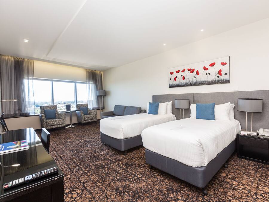 Habitación de hotel con dos camas, un escritorio, un televisor y un amplio ventanal con vistas a la ciudad.