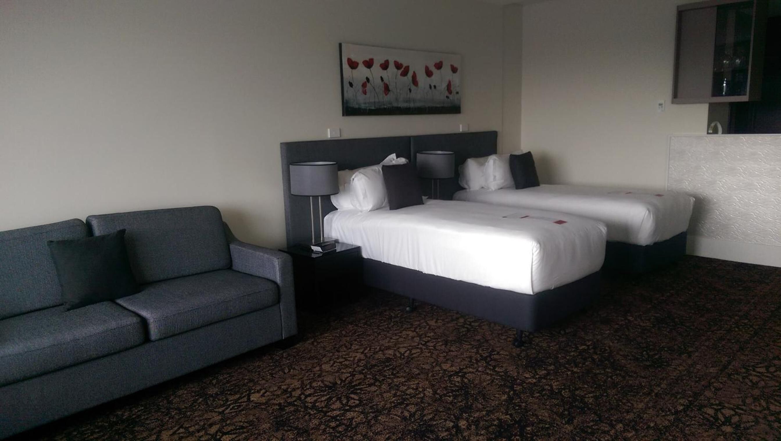 A hotel room with a double bed, a single bed, a grey sofa, and a painting on the wall.