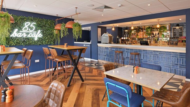 Indretning i en bar og café med trægulv, læder- og metalstole, en bardisk med skrammel og et neonlys på væggen.