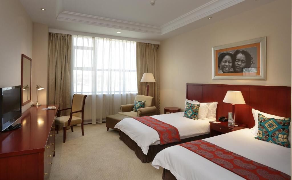 A hotel room with two beds, a desk with a TV, a chair, a lamp, and a window with curtains.
