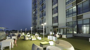 Un espacio al aire libre con mobiliario moderno, un rascacielos al fondo y cielo despejado.