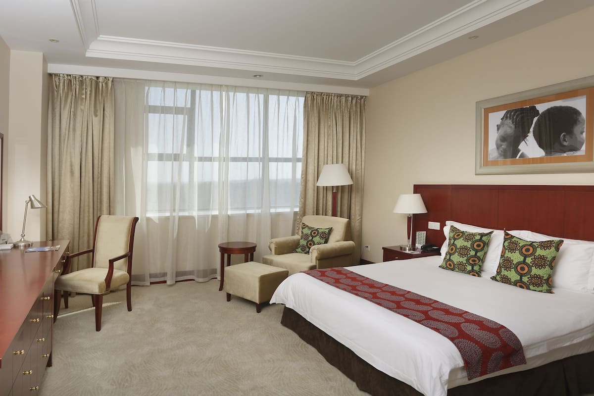 A hotel room with a bed, two armchairs, a desk with a TV, a lamp, and a large window with curtains.