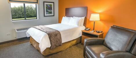 A hotel room with a bed, a nightstand, a lamp, a chair, and a window with a view of trees.