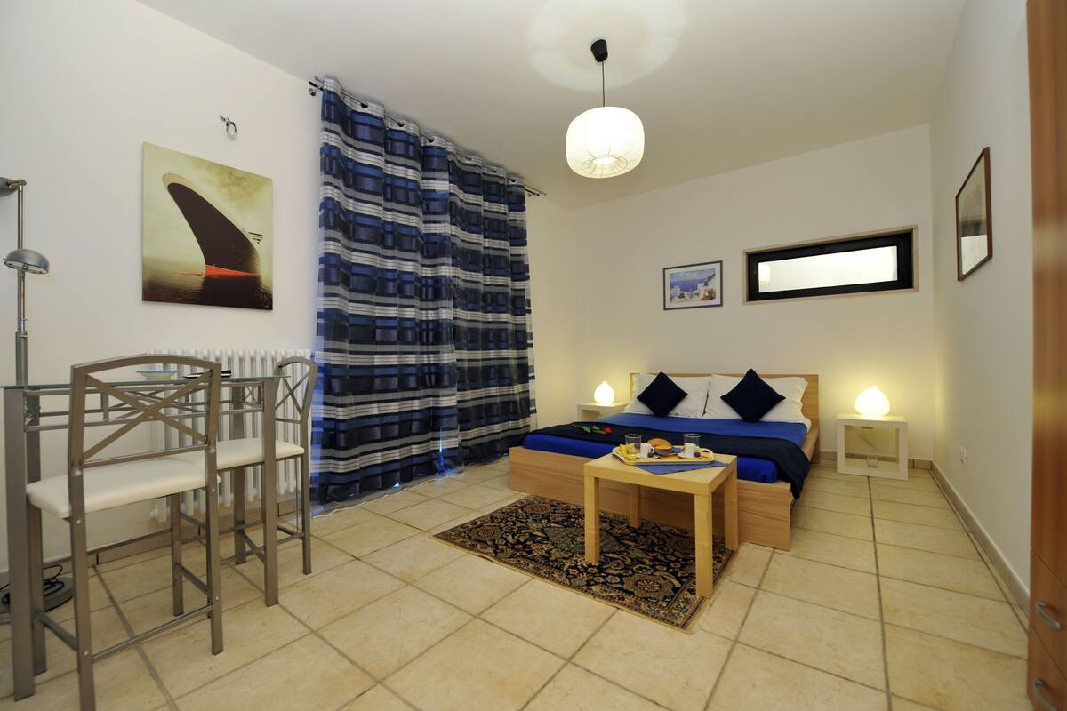A room with a bed, bedside tables, a chair, a table with a tray, a radiator, a curtain, a painting, and a window.