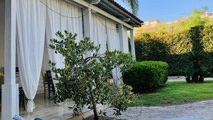 A covered outdoor seating area with a wicker lounge chair, a small tree, and a building with a tiled roof and white curtains. - B&B A Casa di Joy (Lecce)